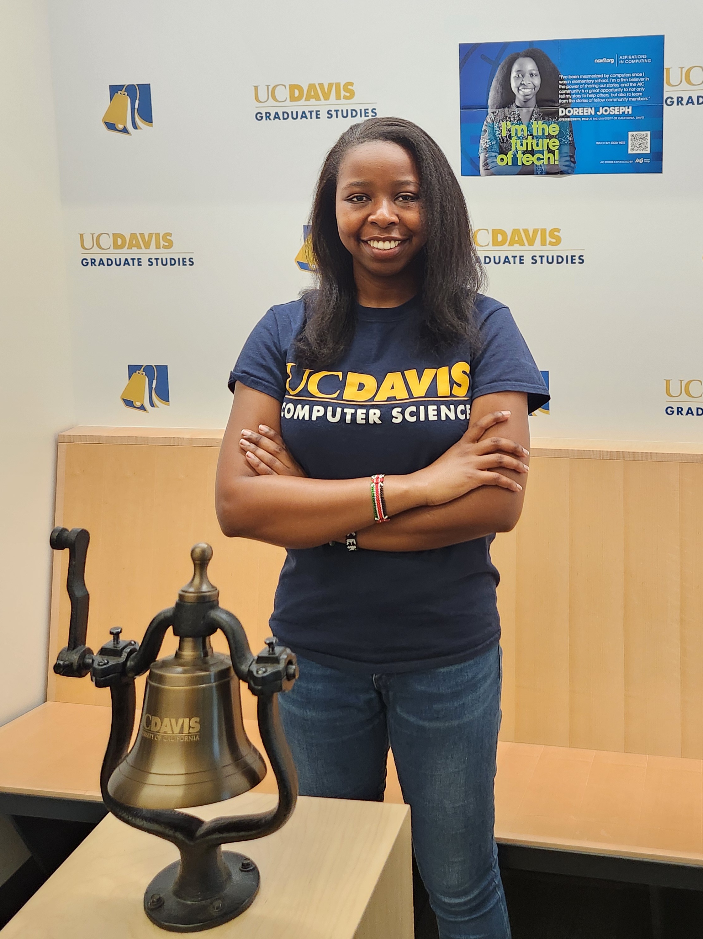 Doreen Joseph stands in Walker Hall next to the hallmark bell that graduate students ring when they finish their Ph.D. Behind Joseph is a poster of herself on the wall.