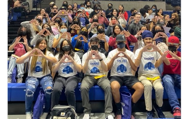 A crowd gathers in an auditorium wearing their CCYLC shirts. In the front row, five students hold up their fingers to make the shape of a heart.