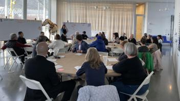 Groups of people sitting at round tables and in conversation with someone speaking near a white board
