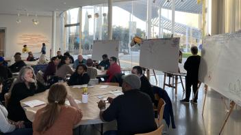 Groups of people sitting at round tables and in conversation with someone speaking near a white board