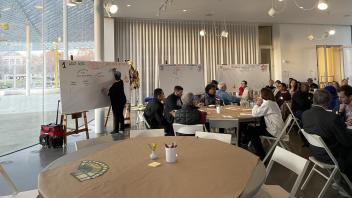 Groups of people sitting at round tables and in conversation with someone speaking near a white board