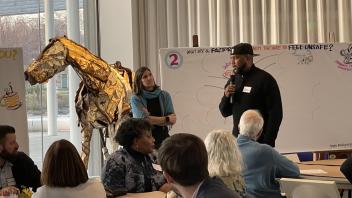 A Black man holds the microphone and speaks to a group sitting at a table