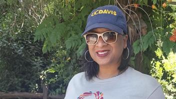 Renetta Tull stands in front of an autumn landscape, wearing sunglasses and decked out in a blue hat and gray t-shirt with the UC Davis Diversity, Equity and Inclusion, logo. 