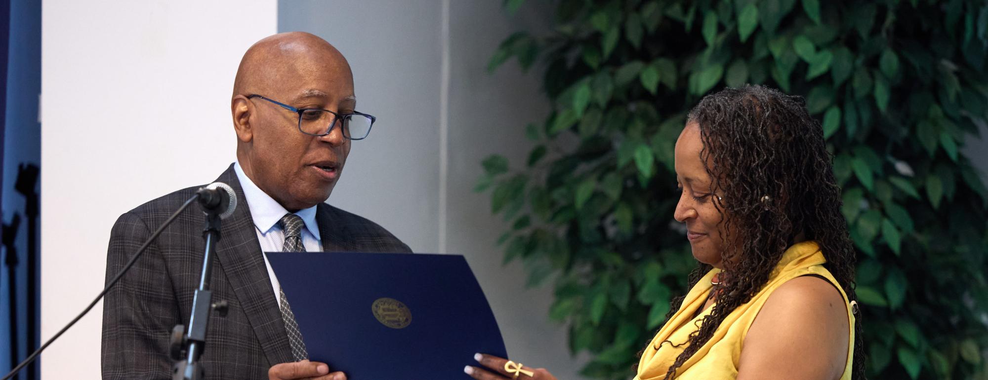 A man in a suit presents a certificate to a woman in a yellow top and patterned skirt at an indoor event.