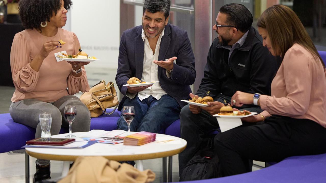 Four adults sitting on purple couches share a light meal and conversation in a casual indoor setting. They are smiling and laughing while holding plates of food, with drinks and brochures on the round table in front of them. The atmosphere appears warm and social.