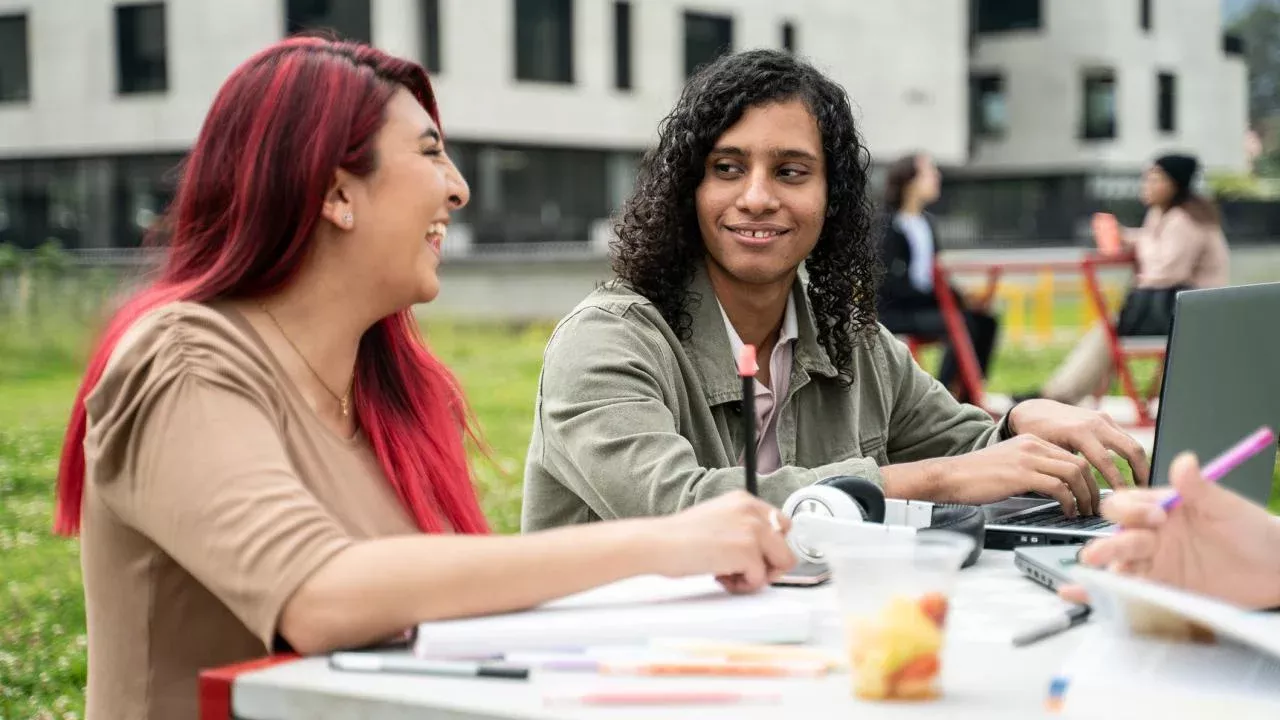 Woman with long hair dyed bright red sits at a table, laughing and engaged in conversation with man with shoulder-length curly hair who is working on a laptop.