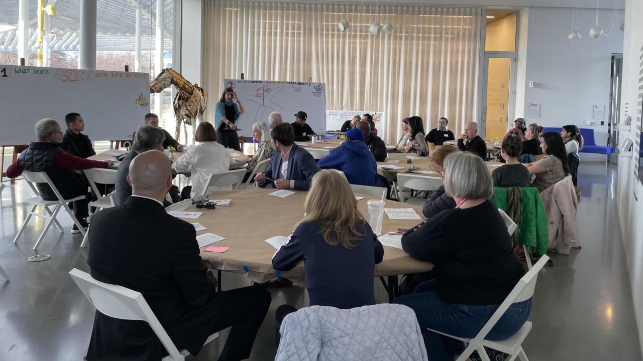 Groups of people sitting at round tables and in conversation with someone speaking near a white board