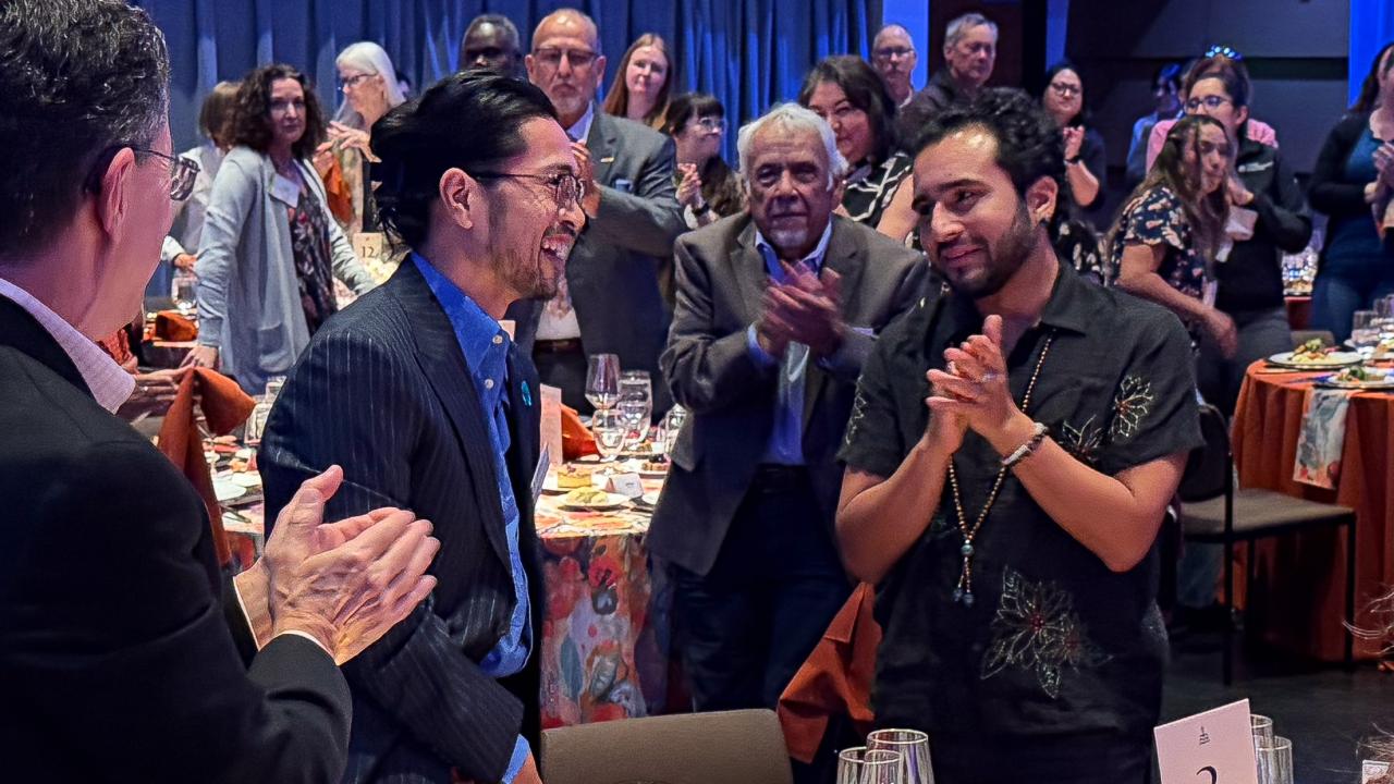 A crowd applauds two men engaged in conversation at a colorful banquet setting.