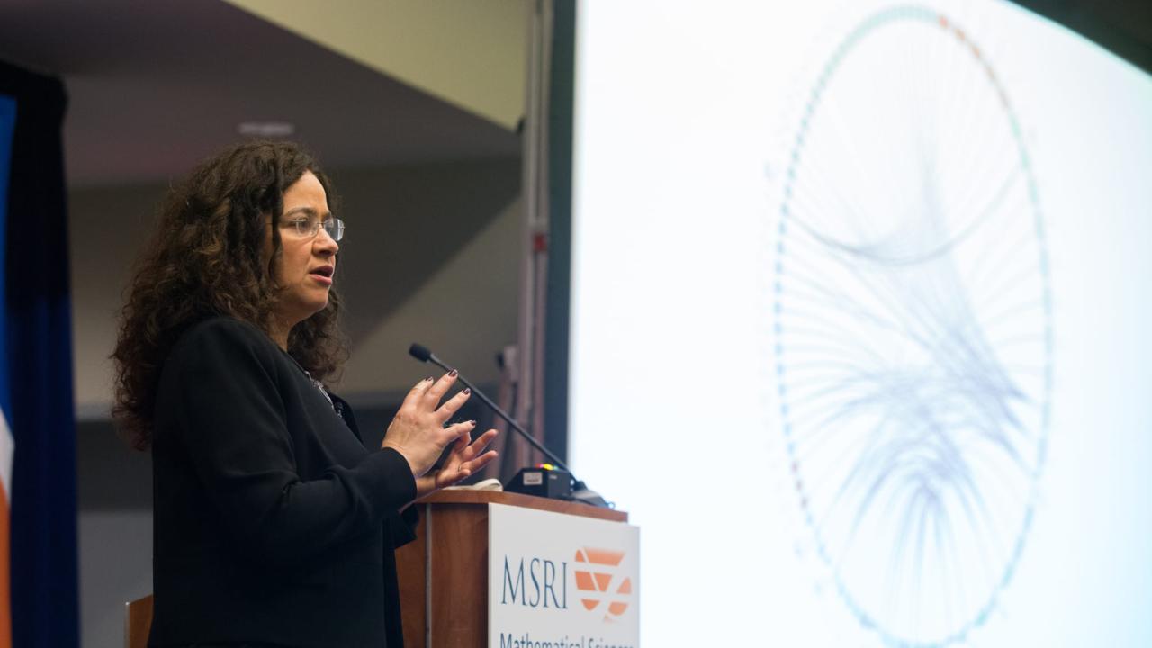 Woman speaking at podium, large circular network graphic projected on screen