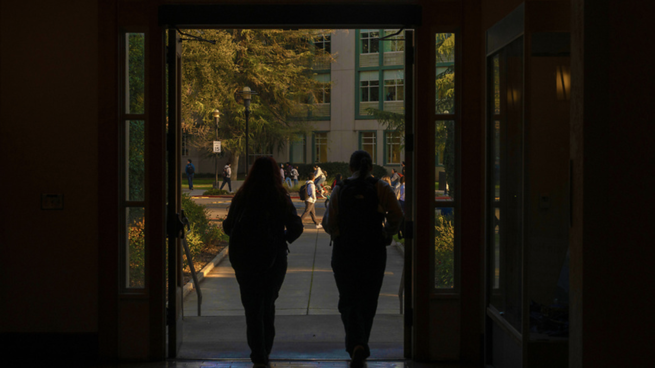 Two people with backpacks walk out of a building into a sunlit campus courtyard, where other students are walking and a multi-story academic building and trees are visible in the background.