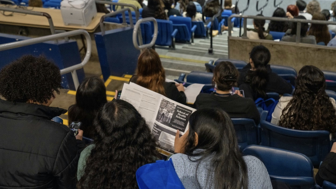 Audience members seated in an auditorium, with one person in the foreground reading a program booklet while others listen to a presentation on stage.