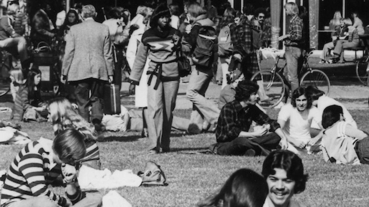 Black and white photo of students gathered on a campus lawn, with buildings and trees in the background.