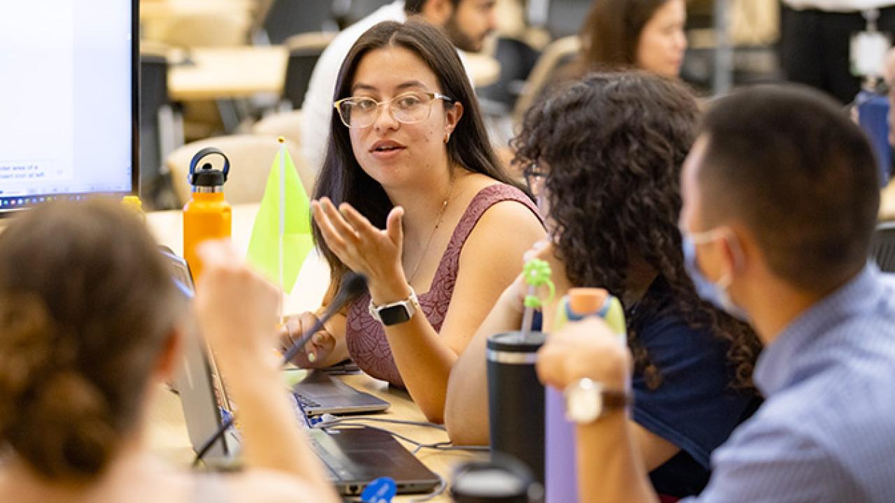 Students sit together at a table in a classroom, engaged in discussion with laptops and notebooks open, while one student gestures as she speaks.