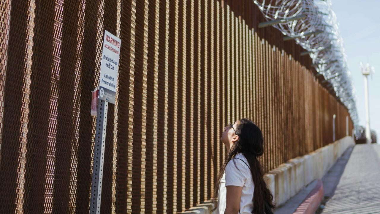 A woman gazes up at a tall border wall topped with barbed wire under a clear sky.
