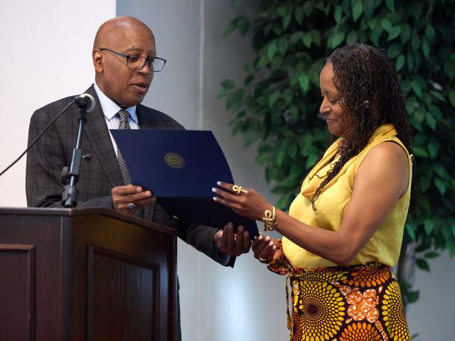 A man in a suit presents a certificate to a woman in a yellow top and patterned skirt at an indoor event.