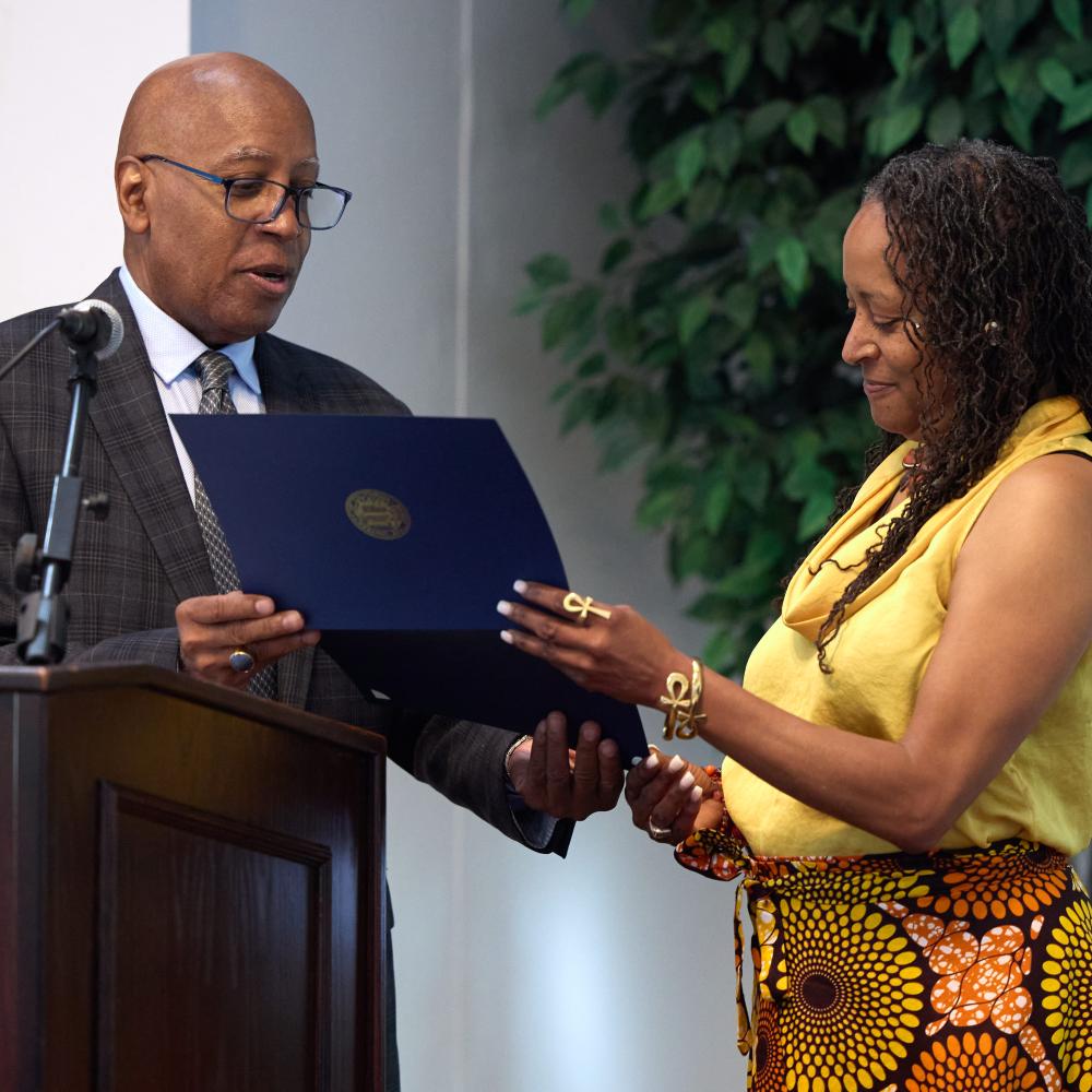 A man in a suit presents a certificate to a woman in a yellow top and patterned skirt at an indoor event.
