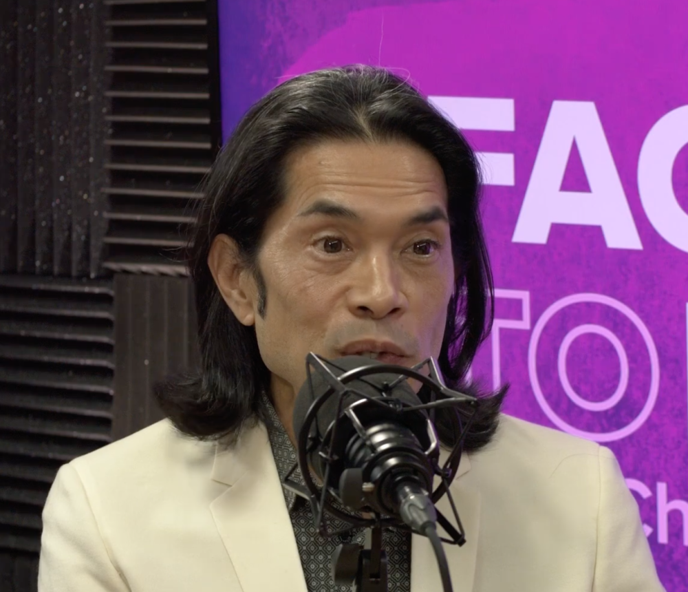 Person speaking into a microphone in front of a purple background with text.