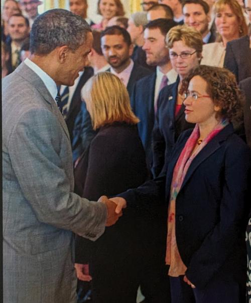 Man in gray suit shakes hands with woman in dark blazer and pink scarf, onlookers behind.