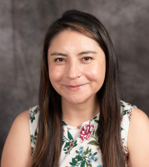 Smiling Marie-Jose-Echeverria with long hair wearing a floral-patterned top, against a gray background.