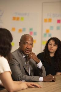 Chancellor Gary S. May seated at a conference table speaking with two students.