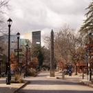 A tree-lined street in downtown Davis, California, featuring the colorful mosaic-covered Davis Needle sculpture in the center, with pedestrians walking on the sidewalk, bicycles parked to the right, and the UC Davis campus buildings visible in the background under a cloudy sky. Bucket Options