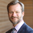 Portrait of a man with sandy blonde hair, a beard and a mustache wearing a blue suit and tie