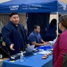 Indoor health fair with a blue UC Davis Health tent and table, with people interacting.