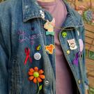 A person cropped from neck to waist wearing a jean jacket with stitching that reads "Queer," buttons that represent UC Davis, a red ribbon, a purple ribbon, a heart, a cat, a daisy, and a button that says "they, them, theirs"  