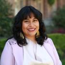 Michelle Vasquez Ruiz  with curly black hair, wearing earrings and a light purple jacket, outdoors.