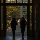 Two people with backpacks walk out of a building into a sunlit campus courtyard, where other students are walking and a multi-story academic building and trees are visible in the background.