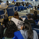Audience members seated in an auditorium, with one person in the foreground reading a program booklet while others listen to a presentation on stage.