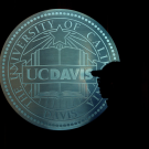 A silhouetted person in profile stands in front of an illuminated University of California, Davis seal, featuring an open book and star, against a dark background.