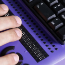 A hand rests on a purple braille display next to a black keyboard.