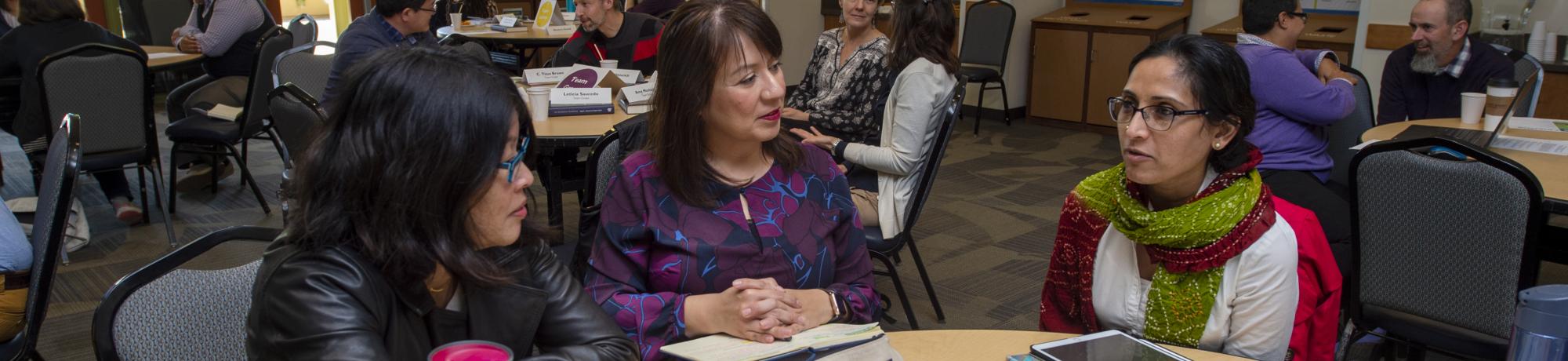 Three women sit around a table engaged in conversation. In the background, other women and men are seated at round tables engaged in coversation.